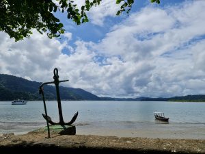 A weathered anchor stands on the shoreline overlooking a calm tropical bay, while small boats rest on the water beneath a sky filled with dramatic clouds