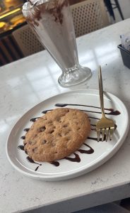  A plate featuring a chocolate chip cookie alongside a tall glass of creamy milkshake.
