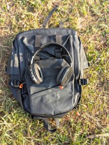 A black backpack is placed on green grass. It has a sleek, modern design, with a front pocket that appears slightly unzipped.
