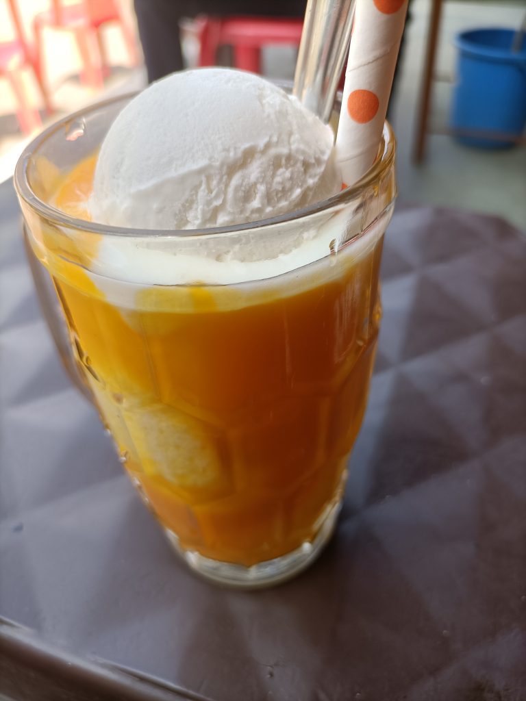 Vanilla ice cream with mango drink served in a tall glass with straws on a brown table.
