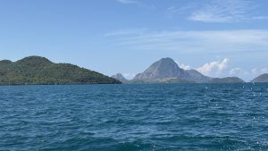Turquoise sea ripples toward lush, forested islands and dramatic mountain peaks beneath a clear blue sky, creating a serene coastal scene.