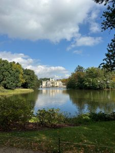 A serene landscape featuring a calm lake surrounded by lush greenery. In the foreground, there are shrubs and small plants with some hints of autumn colors. The water reflects the bright blue sky creating a tranquil atmosphere. In the background, a large, elegant building with large windows can be seen, positioned near the edge of the lake.