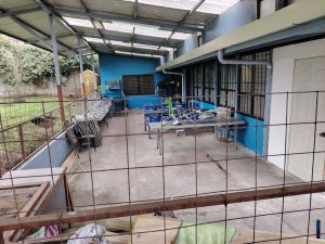 Sheltered outdoor cafetería area with stacked folding chairs, long tables, and and wire mesh security fencing
