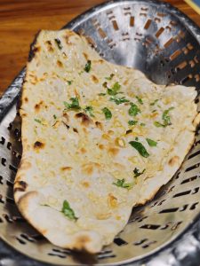 A freshly prepared garlic naan served in a metal basket, topped with chopped garlic, coriander and a light coating of butter. The toasted spots and toppings are clearly visible. Location: Edavannappara, Malappuram, Kerala