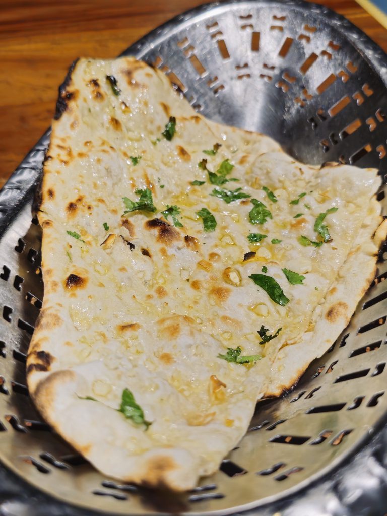 A freshly prepared garlic naan served in a metal basket, topped with chopped garlic, coriander and a light coating of butter. The toasted spots and toppings are clearly visible. Location: Edavannappara, Malappuram, Kerala