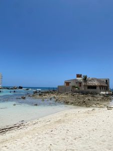 Peaceful tropical beach with clear shallow water, white sand, scattered rocks, and a small rustic seaside house with a traditional thatched roof beneath a bright blue sky.
