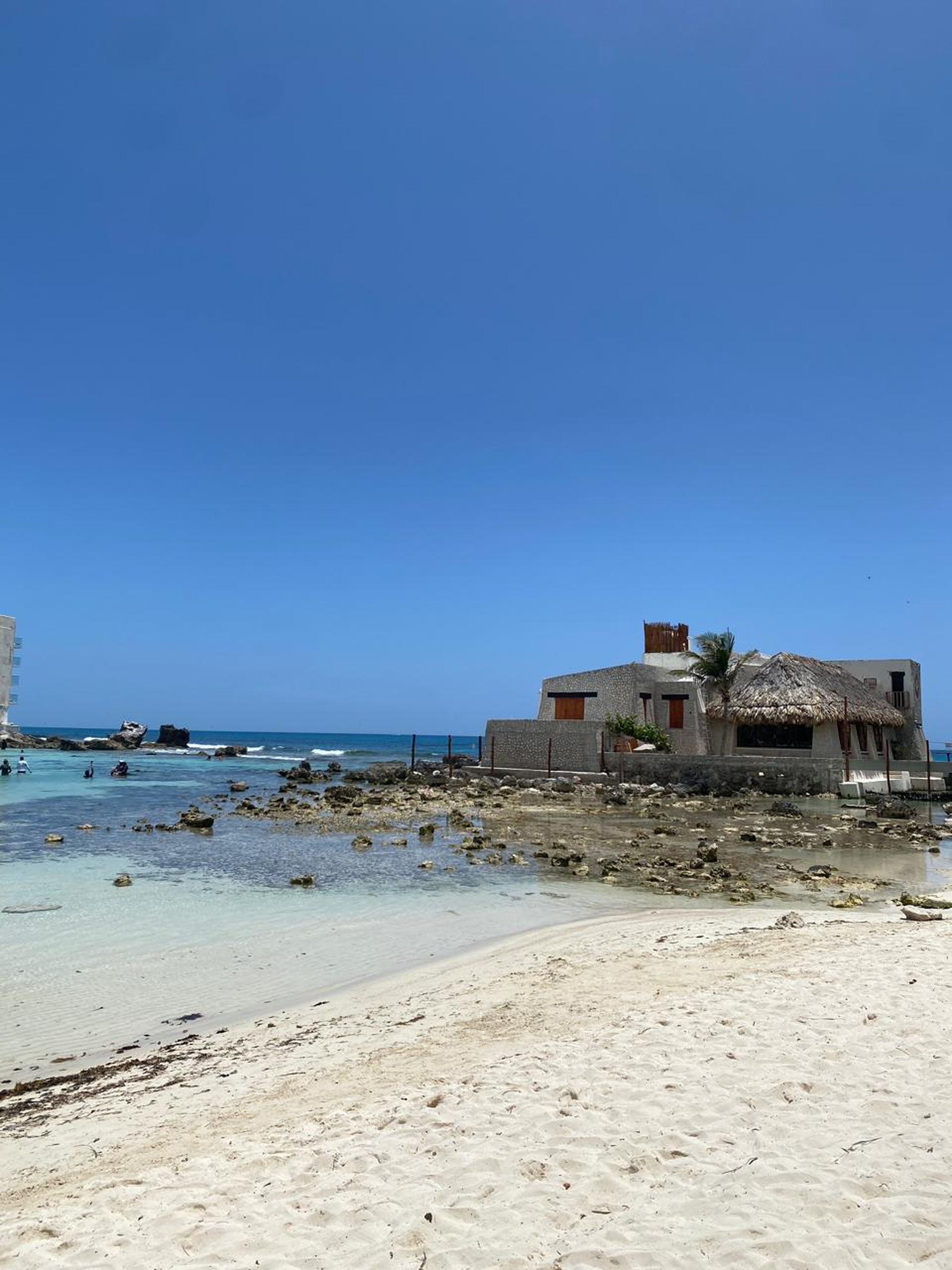 Peaceful tropical beach with clear shallow water, white sand, scattered rocks, and a small rustic seaside house with a traditional thatched roof beneath a bright blue sky.