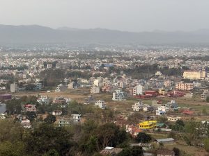 Urban landscape with colorful low-rise buildings, green trees, and distant mountains under a hazy sky.