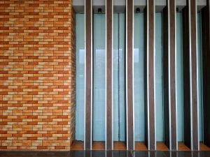 Modern building exterior wall featuring a warm reddish-brown brick tile section on the left and vertical dark wood louvers over frosted glass panels on the right.
