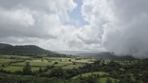 A lush green landscape stretches out under a dramatic sky filled with thick gray clouds. Rolling hills rise in the background, partially obscured by the clouds. In the foreground, fields of tall grass and crops are interspersed with clusters of trees and shrubs.
