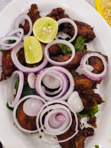 A plate of crispy fried chicken pieces (Chicken 65) is topped with rings of fresh onion, lemon halves, green chili, and curry leaves. The dark fried pieces contrast strongly with the bright garnishes. Captured in Palazhi, Kozhikode, Kerala.