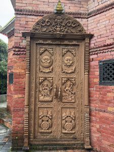 An intricately carved brown wooden double door is set within a red-brick wall. The top arch features detailed relief carvings topped with a small golden emblem. Each door panel depicts traditional relief carvings: a sun and moon face at the top, Hindu deities in the center, and ceremonial sacred pots at the bottom.