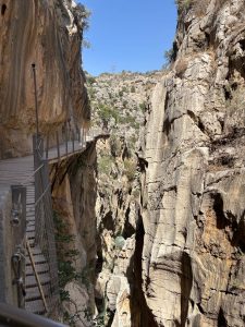 A narrow wooden walkway crosses a rocky canyon with steep cliffs and a river below.