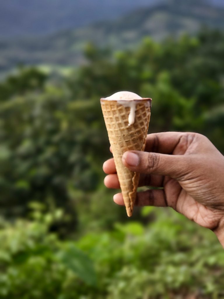A hand holds a cone of vanilla ice cream with a small amount melting over the edge. The green hills and vegetation in the background are softly blurred, creating a strong focus on the ice cream and giving the photograph a pleasant outdoor holiday feeling. Location: Kariyathumpara, Kozhikode, Kerala.