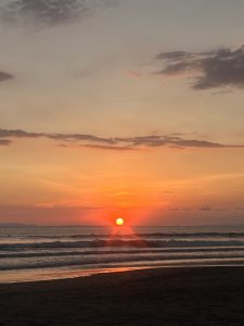 A vibrant sunset over the ocean, where the sun appears as a glowing orange sphere just above the horizon. The sky is a gradient of colors, transitioning from deep orange near the sun to soft blues and grays higher up, with scattered clouds adding texture.