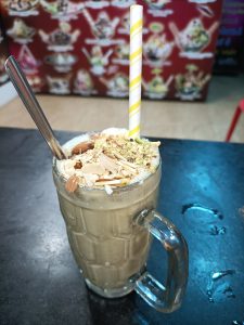 Tall glass of creamy lassi topped with ice cream and nuts, with two straws on a dark table.
