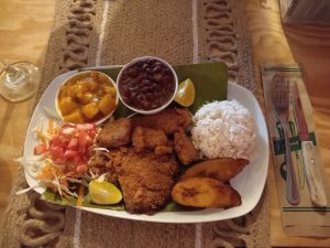 
A plate of breaded fish with white rice, beans, fried ripe plantains, salad, potato hash, and lemon slices.