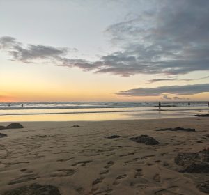 A wide sandy beach stretches out under a colorful sky at sunset, showcasing shades of orange and soft pink. Gentle waves lap at the shore, leaving behind glistening wet sand. In the distance, a few figures can be seen wading in the water.