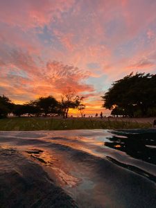A tranquil scene capturing a colorful sunset. The sky is filled with layers of pink, orange, and purple clouds, creating a vivid gradient as the sun sets on the horizon. In the foreground, the surface of a body of water reflects these warm hues, rippling gently.