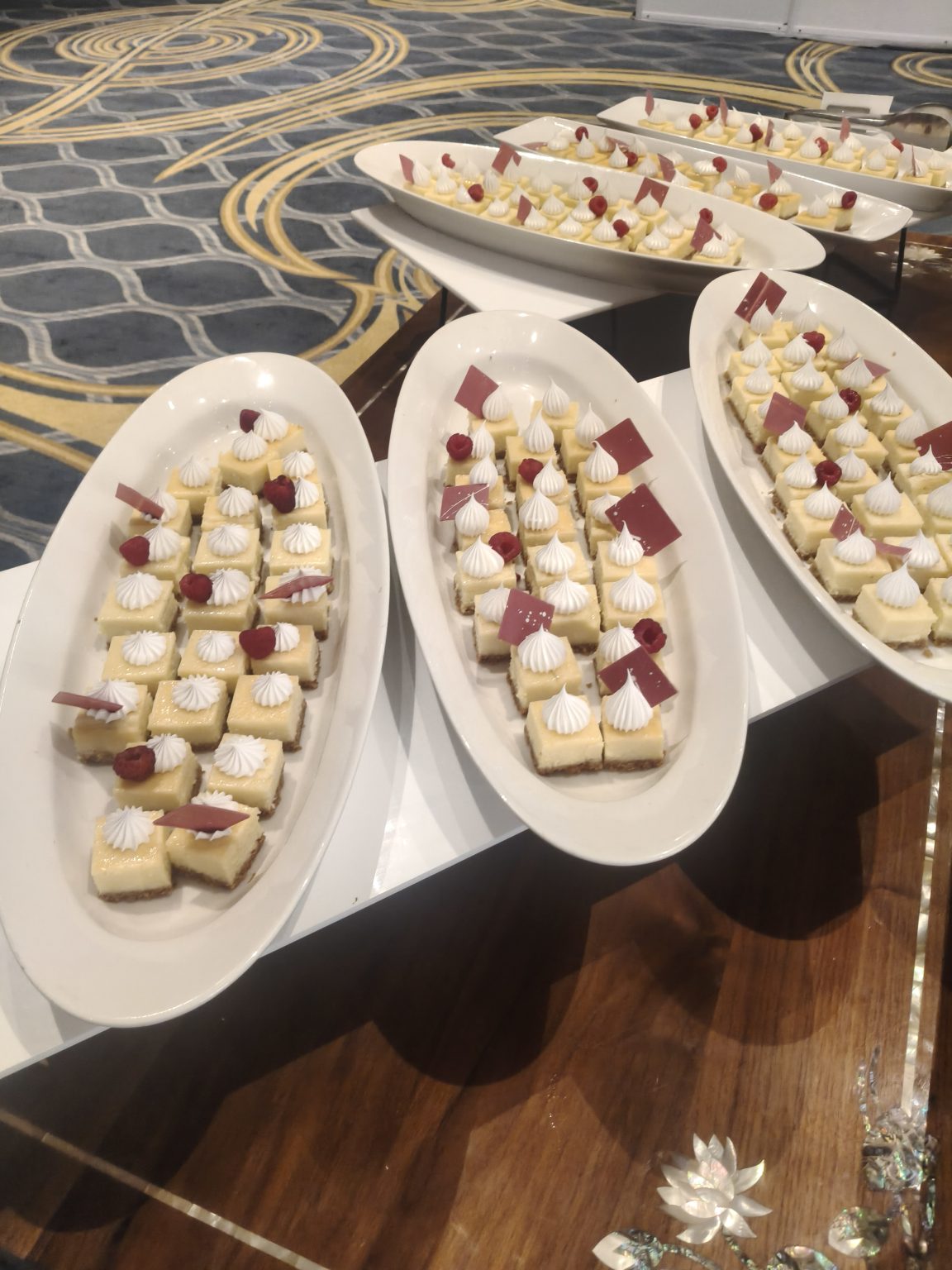 A platter of small square desserts topped with white meringue swirls and red raspberry garnish. Some pieces have small chocolate decorations.