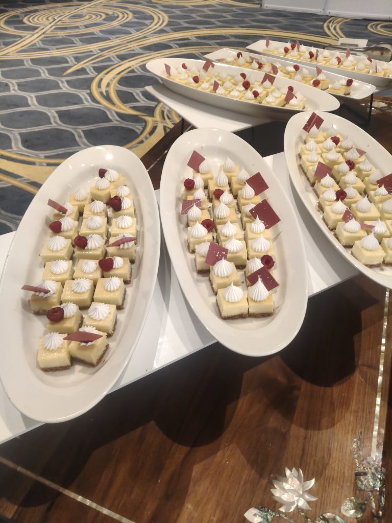 A platter of small square desserts topped with white meringue swirls and red raspberry garnish. Some pieces have small chocolate decorations.