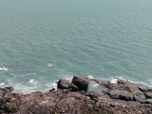High-angle view of greenish-blue ocean waves gently crashing against dark, rugged coastal rock formations in the foreground, expanding out to a vast horizon under bright daylight.