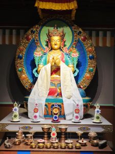A richly decorated Buddhist sculpture sits in a shrine-like setting, surrounded by colorful ornaments, flowers, lamps and traditional offerings. The golden figure and bright blue, green and red details create a striking display of South Asian religious art. Captured at Chhatrapati Shivaji Maharaj Vastu Sangrahalaya, Mumbai, Maharashtra.