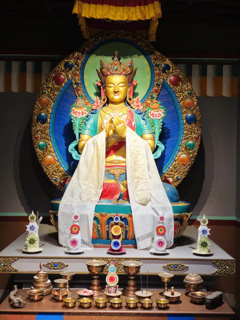 A richly decorated Buddhist sculpture sits in a shrine-like setting, surrounded by colorful ornaments, flowers, lamps and traditional offerings. The golden figure and bright blue, green and red details create a striking display of South Asian religious art. Captured at Chhatrapati Shivaji Maharaj Vastu Sangrahalaya, Mumbai, Maharashtra.