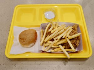A simple meal served on a bright yellow tray with crispy fried chicken, golden French fries, a soft bread roll, and a cup of creamy dip. The colorful presentation makes it look inviting. Captured in Palazhi, Kozhikode, Kerala.