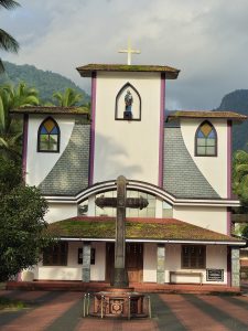 A beautiful church stands against the green hills of Kariyathumpara. The white building has a tall central tower, colorful windows, tiled roofs, and a large cross in front. Coconut trees and forested hills create a peaceful natural setting around the church. Location: Kariyathumpara, Kozhikode, Kerala.