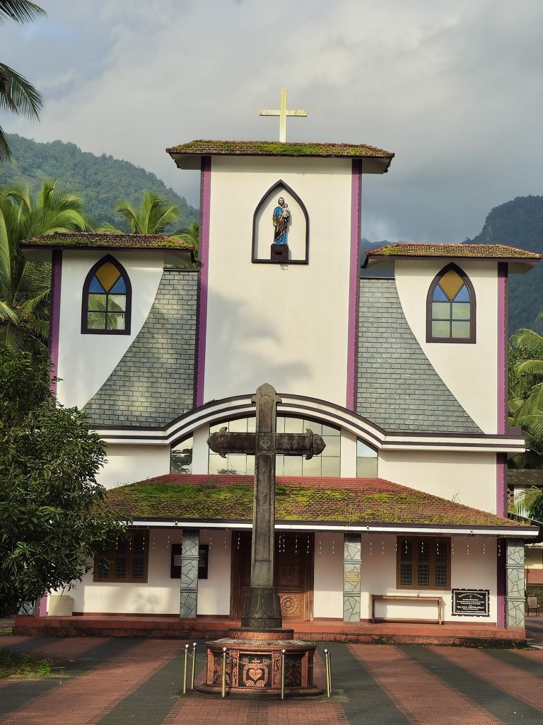 A beautiful church stands against the green hills of Kariyathumpara. The white building has a tall central tower, colorful windows, tiled roofs, and a large cross in front. Coconut trees and forested hills create a peaceful natural setting around the church. Location: Kariyathumpara, Kozhikode, Kerala.