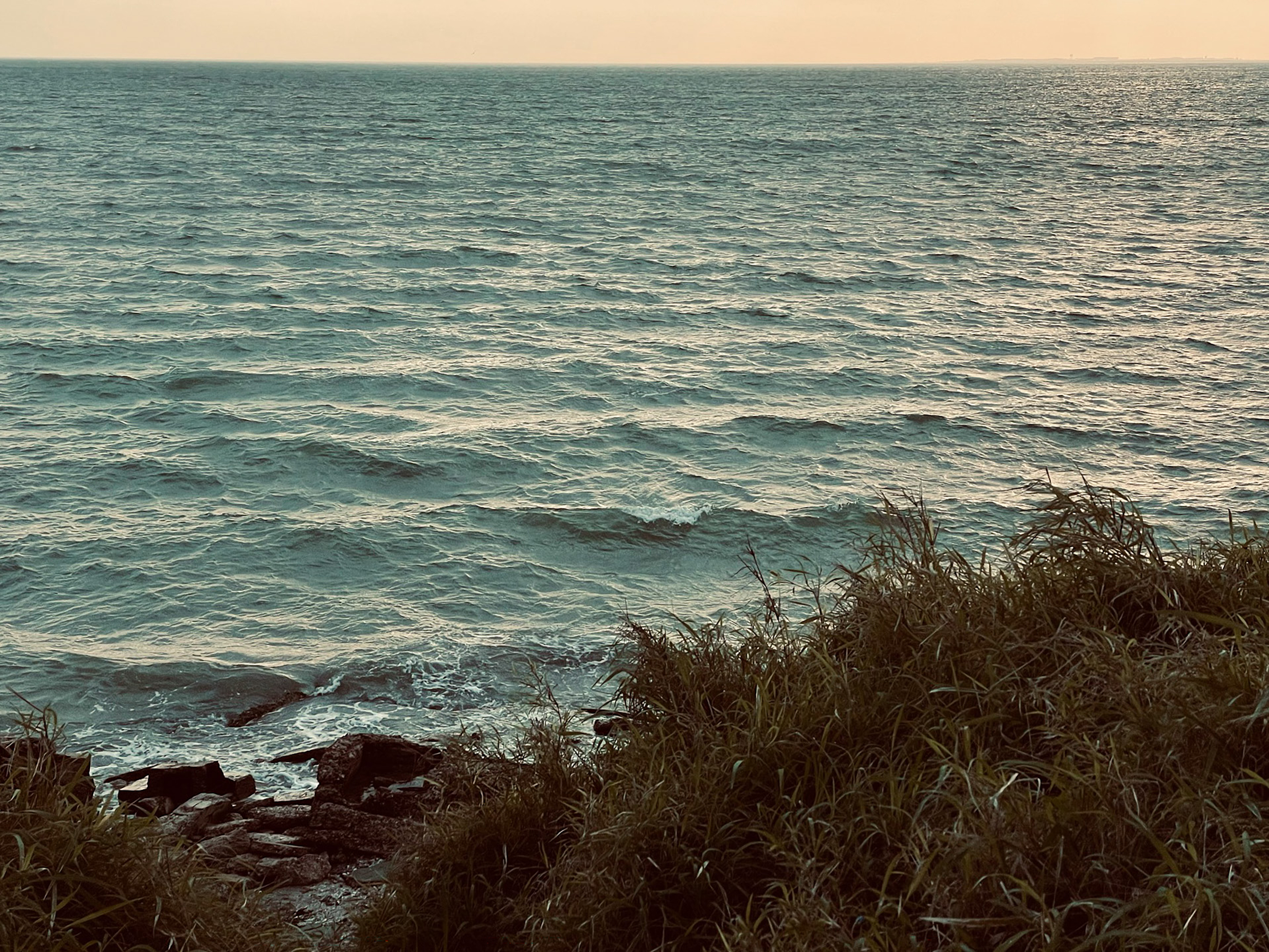 Ocean waves viewed from a grassy, rocky shoreline beneath a warm, hazy sky.
