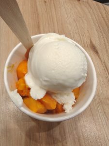 A paper cup sits on a wooden surface, filled with a scoop of white, creamy ice cream atop bright orange chunks of mango.