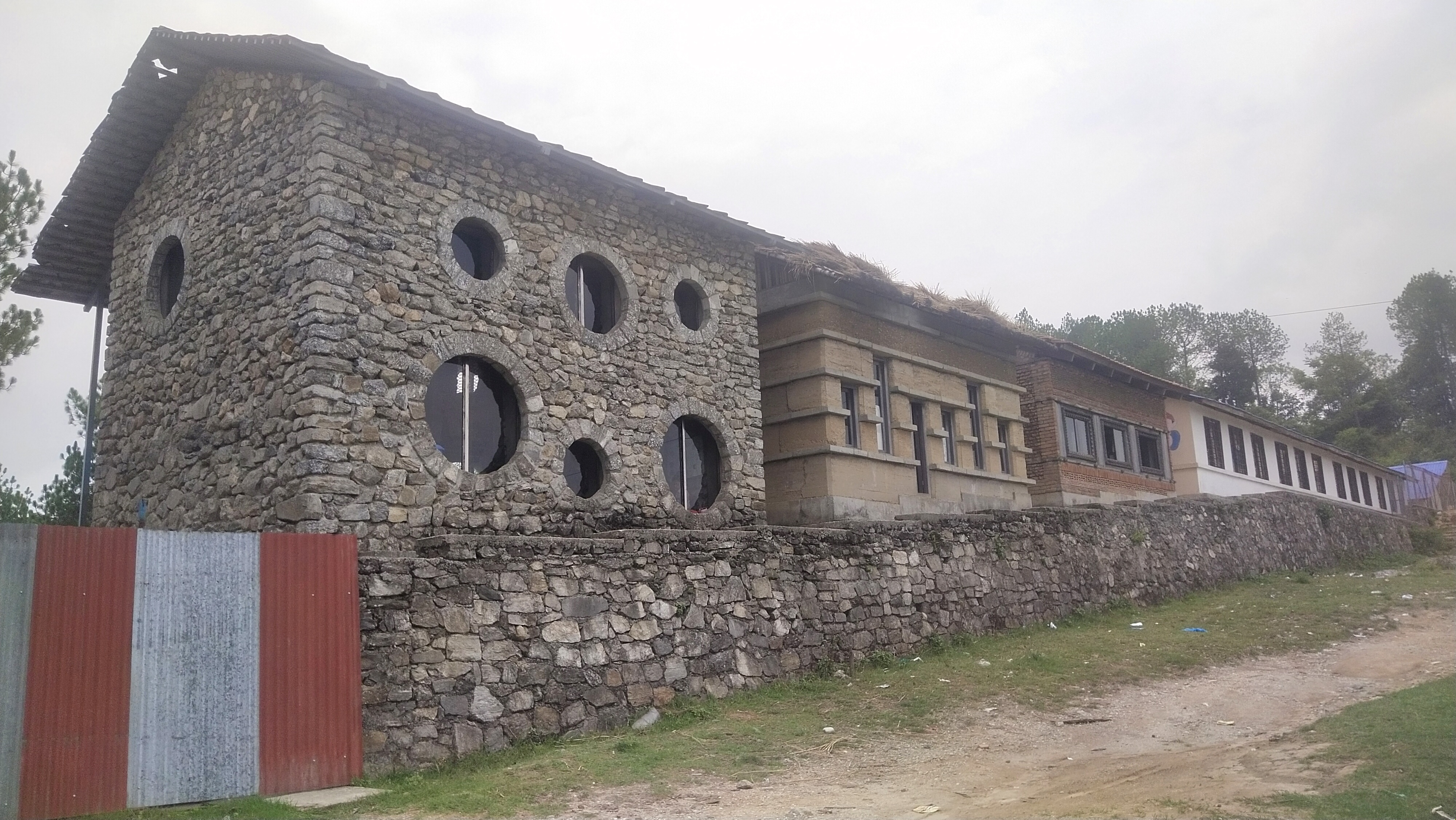 Four rustic buildings in a row on a slight incline, each built a different medium and style, a two storey stone structure with circular windows under a corrugated roof is the most prominent. A metal corrugated fence to the left and a stone wall runs in front of all of the buildings. A litter strewn grass and dirt track in the foreground.