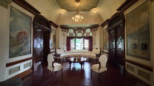 Elegant historic sitting room with dark wood paneling, arched doors, landscape murals, antique chairs and table, and a bright window.