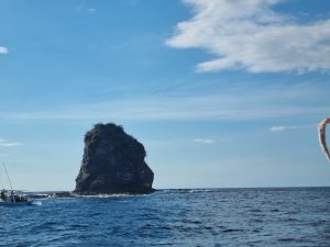 Large rock formation rising from the sea, surrounded by deep blue water, gentle waves, and a small boat nearby.