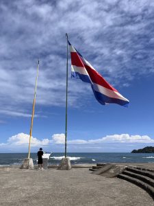 Costa Rican flag and a yellow flag displayed on flagpoles beside an informational sign overlooking the coastline. The ocean, a small green hill, and a bright blue sky with scattered clouds complete the coastal landscape