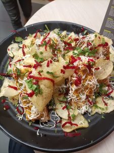A black plate is piled high with a colorful and textured arrangement of snacks. Prominent are crispy potato chips, some whole and others broken into smaller pieces.
