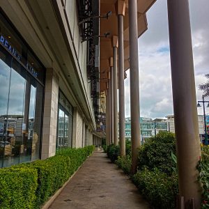 A covered walkway lined with tall pillars, glass store fronts, and lush green plants.