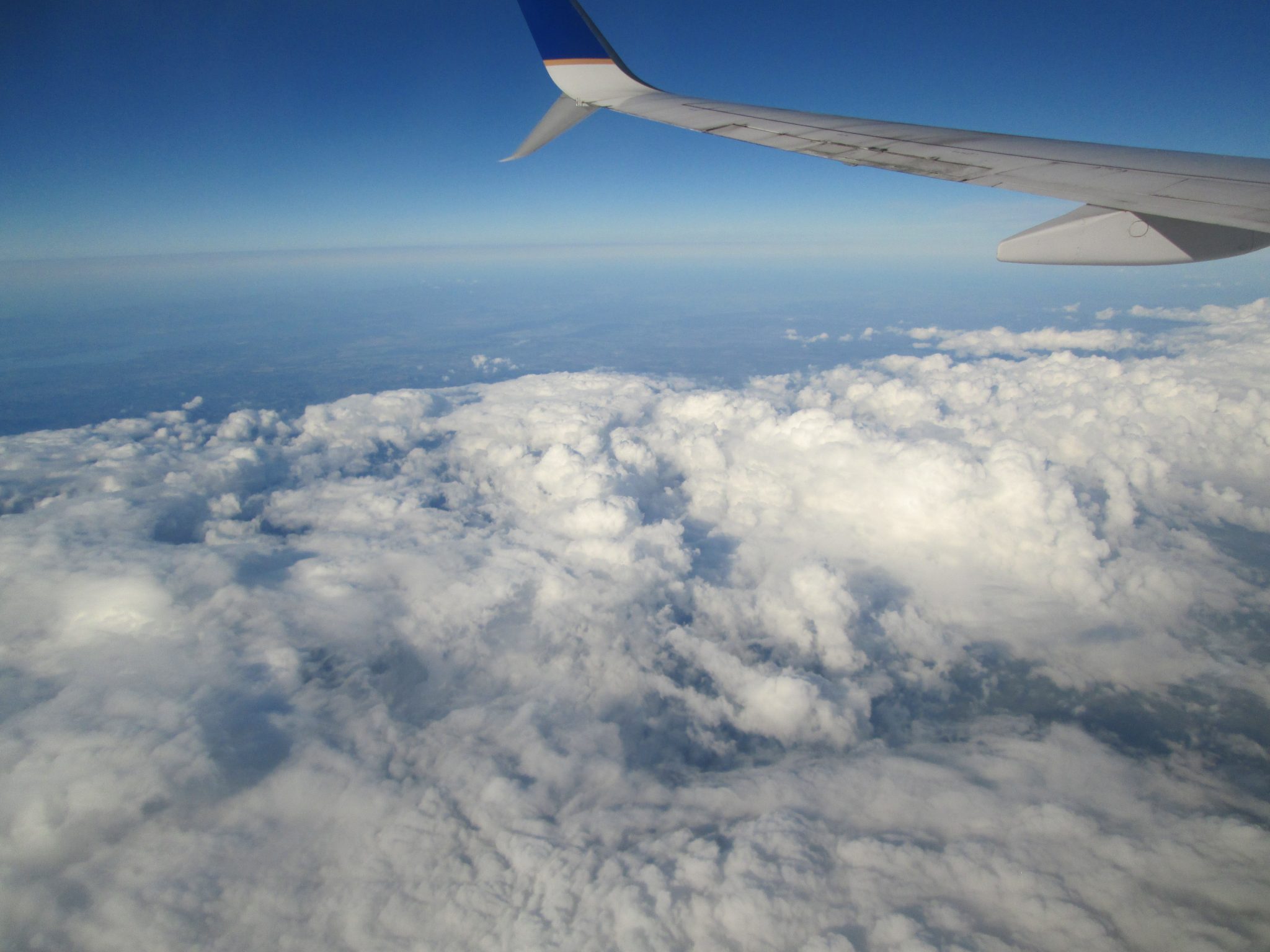 View from an airplane window showing the wing extending over a blanket of white clouds under a clear blue sky. The horizon stretches into the distance, creating scene high above the landscape