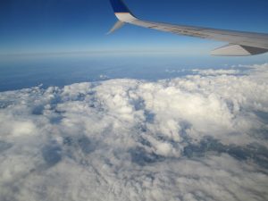 View from an airplane window showing the wing extending over a blanket of white clouds under a clear blue sky. The horizon stretches into the distance, creating scene high above the landscape