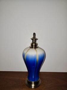 A decorative ceramic bottle with a rounded body features a rich blue glaze fading into pale cream near the top. A metallic lid and base add contrast against the plain background. Captured in Chalappuram, Kozhikode, Kerala.