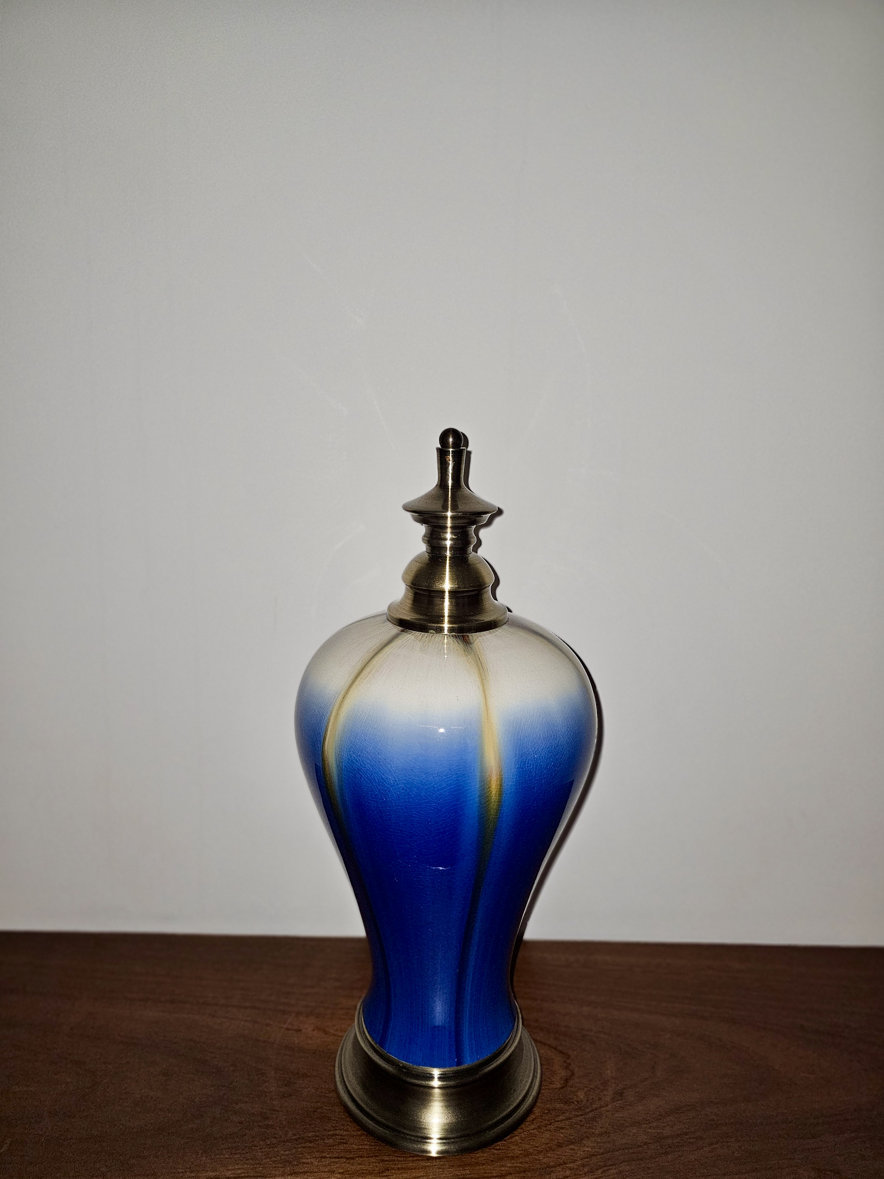 A decorative ceramic bottle with a rounded body features a rich blue glaze fading into pale cream near the top. A metallic lid and base add contrast against the plain background. Captured in Chalappuram, Kozhikode, Kerala.