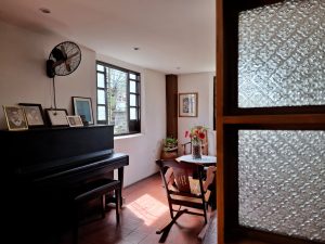 Antique Filipino living room with white walls, terracotta-colored tiles, a piano, an open window, and a wooden rocking chair.