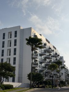 White multi-story residential building with rows of rounded balconies, palm trees, landscaped grounds, and a golf cart parked along the street under a partly cloudy sky.