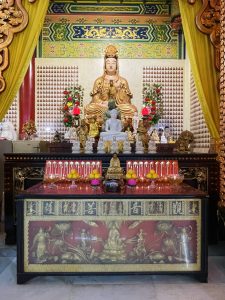 A beautifully decorated Buddhist shrine features a large golden seated statue surrounded by smaller statues, flowers, lamps, and offerings. Bright red, gold, and yellow details add warmth and richness to the scene. Captured at Thean Hou Temple, Kuala Lumpur, Malaysia.