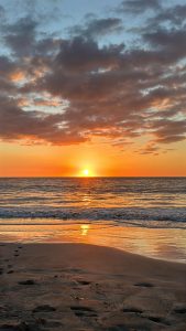 A vibrant sunset over a calm ocean creates a picturesque scene. The sun appears as a bright orange sphere descending toward the horizon, surrounded by colorful clouds ranging from soft pinks to deep oranges. 