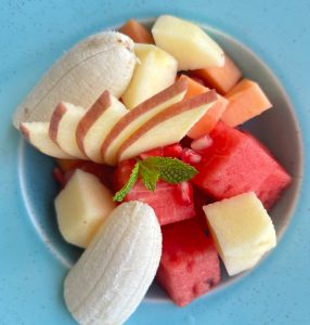 A light blue plate holds a colorful assortment of fruit. The fruits include bright red watermelon cubes, orange cantaloupe pieces, and pale yellow honeydew melon squares
