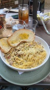 A plate of creamy spaghetti carbonara topped with grated cheese and herbs, served with slices of garlic bread, alongside glasses of iced tea and water on an outdoor wooden table.