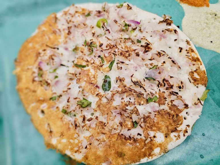 A crispy Uttapam (a type of dosa, commonly known as a South Indian savory pancake or an Indian vegetable pancake) topped with chopped onions, green chili, and herbs is served on a turquoise plate. The golden-brown surface and colorful toppings create an inviting close-up food photograph. Captured in Palazhi, Kozhikode, Kerala.